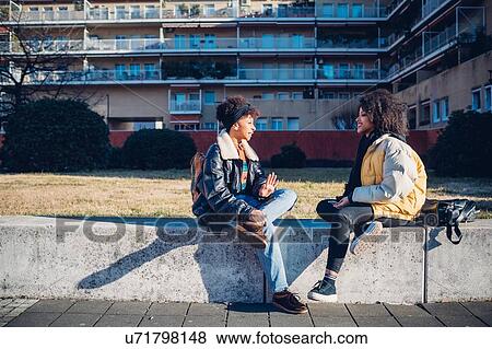 Stock Photo - Two cool young female friends sitting on urban wall chatting. Fotosearch