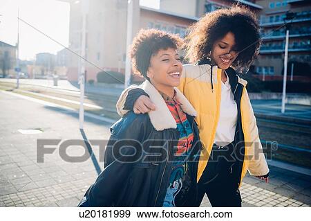 Stock Photo - Two cool young female friends strolling on sunlit urban sidewalk. Fotosearch