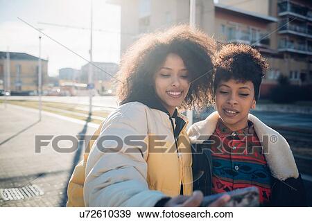 Stock Photo - Two cool young female friends taking smartphone selfie on urban sidewalk. Fotosearch