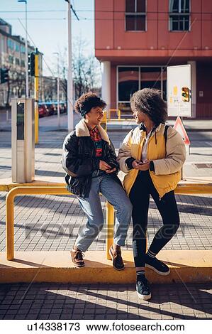 Stock Image - Two cool young women chatting at city tram station. Fotosearch