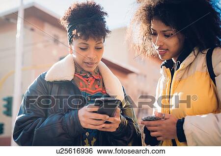 Stock Photograph - Two cool young women looking at smartphone on urban sidewalk. Fotosearch