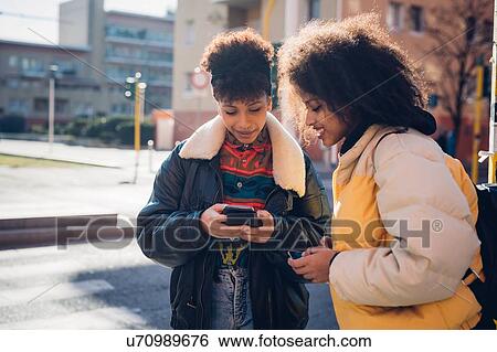 Stock Photograph - Two cool young women looking at smartphone on urban sidewalk. Fotosearch