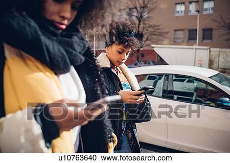 Two cool young women strolling and looking at smartphone on urban sidewalk, sideview View Large Photo Image Stock Image - Two cool young women strolling and looking at smartphone on urban sidewalk, sideview. Fotosearch
