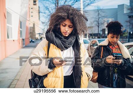 Two cool young women strolling and looking at smartphone on urban sidewalk View Large Photo Image Stock Photograph - Two cool young women strolling and looking at smartphone on urban sidewalk. Fotosearch
