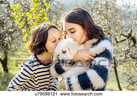 Stock Image - Two girls kissing a cute golden retriever puppy in orchard, Scandicci, Tuscany, Italy. Fotosearch