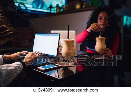 Two women at cafe table drinking smoothie and typing on laptop View Large Photo Image Stock Image - Two women at cafe table drinking smoothie and typing on laptop. Fotosearch