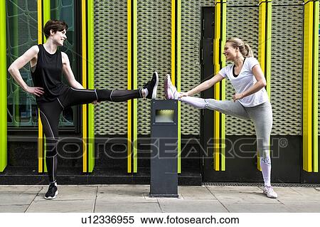 Two young female runners stretching legs on city sidewalk, full length View Large Photo Image Stock Photography - Two young female runners stretching legs on city sidewalk, full length. Fotosearch