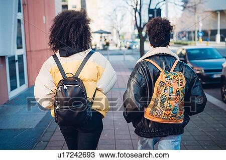 Two young women carrying backpacks strolling on urban sidewalk, rear view View Large Photo Image Stock Image - Two young women carrying backpacks strolling on urban sidewalk, rear view. Fotosearch
