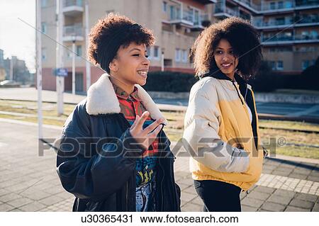 Stock Image - Two young women walking and talking on urban sidewalk. Fotosearch