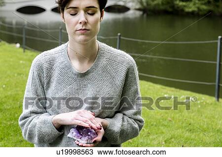 Stock Image - Woman practising Reiki on grass lawn by river, London, UK. Fotosearch