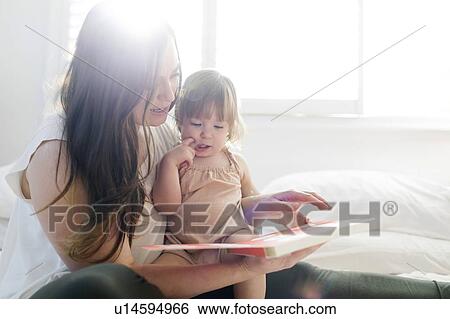 Stock Photograph - Woman reading book to her daughter on bed. Fotosearch