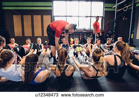 Stock Image - Women training in gym with male trainers, sitting on floor with legs raised. Fotosearch