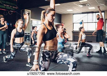 Stock Image - Women training in gym with male trainer, lifting kettle bells with legs outstretched. Fotosearch