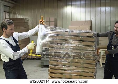 Picture - Workers wrapping stack of cardboard boxes with shrink film in factory. Fotosearch