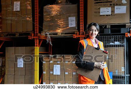 Stock Image - Young female factory worker with clipboard in warehouse, portrait. Fotosearch
