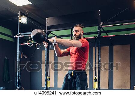 Stock Image - Young man training in gym, lifting kettle bell. Fotosearch