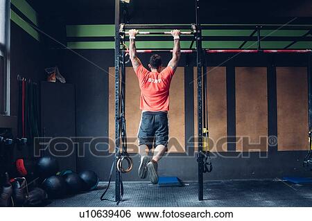 Stock Photograph - Young man training in gym, swinging on exercise bar, rear view. Fotosearch