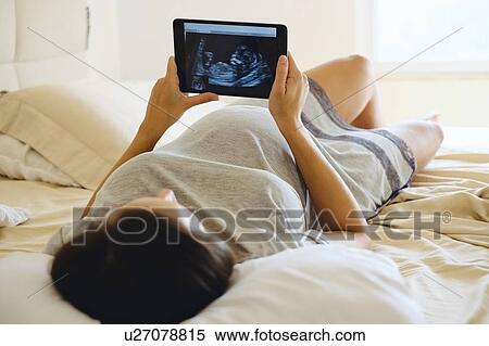 Young pregnant woman lying on bed looking at medical baby scan on digital tablet View Large Photo Image Stock Photography - Young pregnant woman lying on bed looking at medical baby scan on digital tablet. Fotosearch