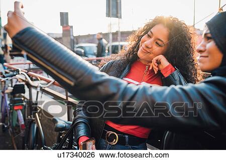 Stock Photograph - Young woman and friend taking selfie in city. Fotosearch