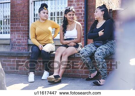 Stock Photo - Young woman and her teenage sisters sitting on wall talking. Fotosearch