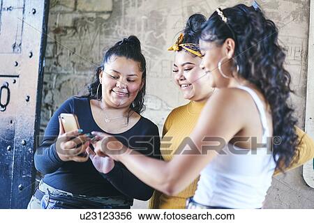 Stock Photograph - Young woman and her teenage sisters looking at smartphone in building entrance. Fotosearch
