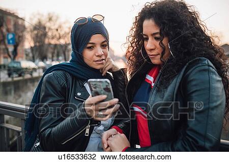 Stock Image - Young woman in hijab and best friend looking at smartphone on canal bridge in city. Fotosearch