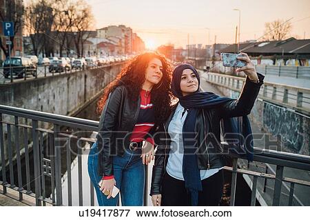 Young woman in hijab and best friend taking selfie on canal bridge in city at sunset View Large Photo Image Stock Photo - Young woman in hijab and best friend taking selfie on canal bridge in city at sunset. Fotosearch