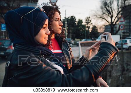 Stock Photo - Young woman in hijab and best friend photographing from canal bridge in city. Fotosearch