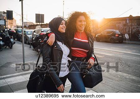 Stock Photography - Young woman in hijab and best friend walking and talking in city at sunset. Fotosearch