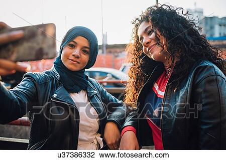 Stock Image - Young woman in hijab and friend posing for selfie in city. Fotosearch