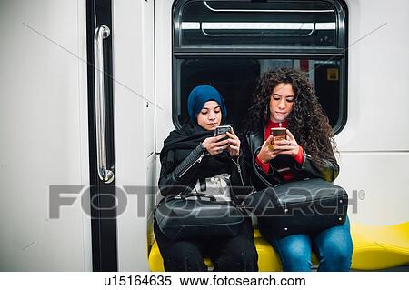 Young woman in hijab and friend sitting on subway train looking at smartphones View Large Photo Image Stock Photography - Young woman in hijab and friend sitting on subway train looking at smartphones. Fotosearch