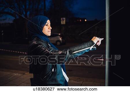Stock Photo - Young woman in hijab using illuminated touchscreen at city bus stop at night. Fotosearch
