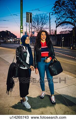 Stock Image - Young woman in hijab with best friend on city sidewalk at dusk, full length. Fotosearch
