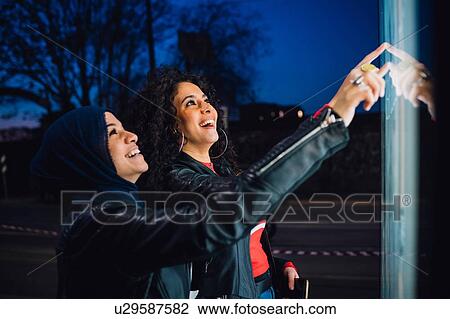 Stock Image - Young woman in hijab with best friend using illuminated touchscreen at city bus stop at night. Fotosearch