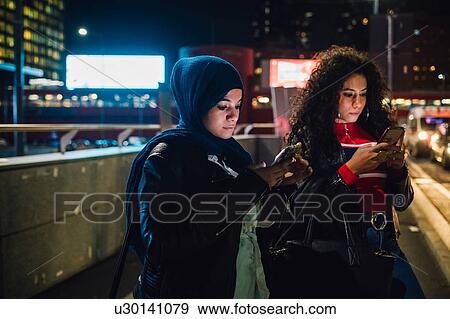 Young woman in hijab with friend looking at smartphones in city at night View Large Photo Image Stock Photo - Young woman in hijab with friend looking at smartphones in city at night. Fotosearch
