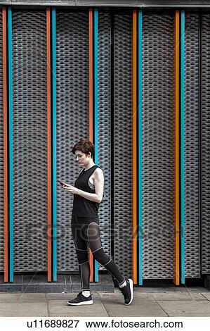 Stock Photo - Young woman in sports clothing looking at smartphone on city sidewalk. Fotosearch