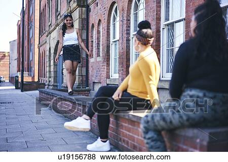 Stock Photo - Young woman walking along wall watched by teenage sisters. Fotosearch
