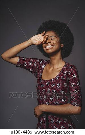 Stock Photography - Young woman with afro hair covering eye and smiling, grey background. Fotosearch