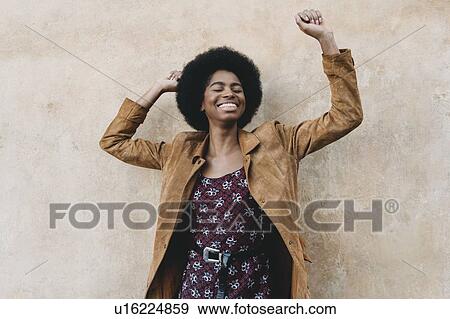 Young woman with afro hair feeling excited against stone wall View Large Photo Image Stock Photo - Young woman with afro hair feeling excited against stone wall. Fotosearch
