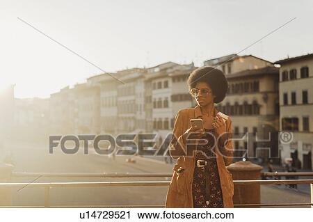 Stock Image - Young woman with afro hair having hot drink, using smartphone in city, Florence, Toscana, Italy. Fotosearch
