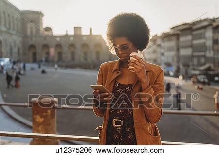 Young woman with afro hair having hot drink, using smartphone in city, Florence, Toscana, Italy View Large Photo Image Stock Photograph - Young woman with afro hair having hot drink, using smartphone in city, Florence, Toscana, Italy. Fotosearch