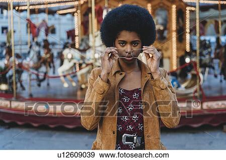 Stock Photo - Young woman with afro hair putting on spectacles in front of carousel. Fotosearch