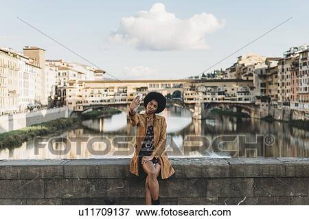 Stock Photo - Young woman with afro hair taking selfie on bridge, Florence, Toscana, Italy. Fotosearch