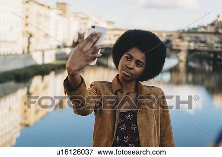 Stock Photo - Young woman with afro hair taking selfie by canal, Florence, Toscana, Italy. Fotosearch