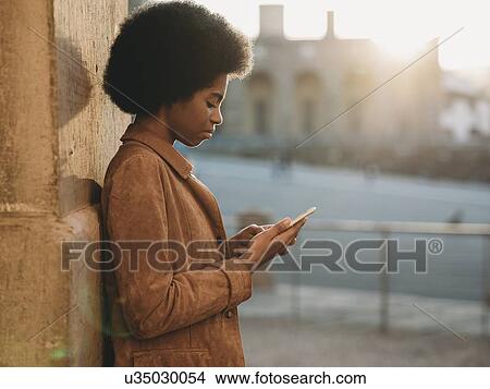 Picture - Young woman with afro hair using smartphone, leaning against stone wall, Florence, Toscana, Italy. Fotosearch