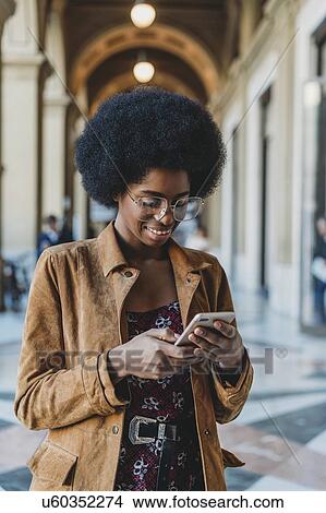 Picture - Young woman with afro hair using smartphone in building corridor. Fotosearch