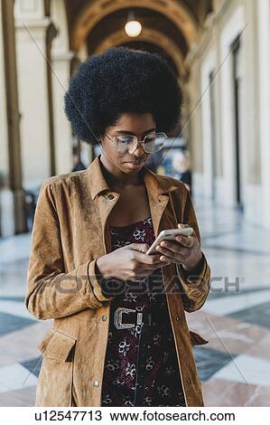 Stock Image - Young woman with afro hair using smartphone in building corridor. Fotosearch