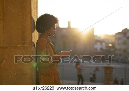 Young woman with afro hair using smartphone, leaning against stone wall, Florence, Toscana, Italy View Large Photo Image Stock Image - Young woman with afro hair using smartphone, leaning against stone wall, Florence, Toscana, Italy. Fotosearch