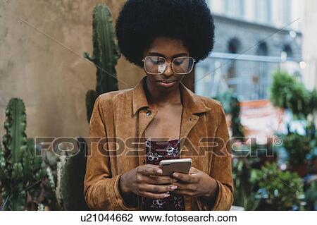 Stock Image - Young woman with afro hair using smartphone in city. Fotosearch