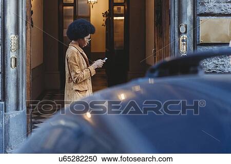 Stock Image - Young woman with afro hair using smartphone in front of building. Fotosearch